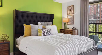 Cozy bedroom at AMLI 900 with green accent wall, tufted headboard, bedside lamps, and a private balcony showing park and skyline views in Chicago
