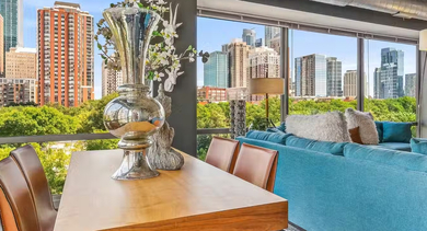 Corner living room at AMLI 900 with large windows framing Chicago skyline views, teal sectional sofa, dining table, and modern industrial ceiling accents