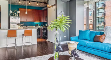 Corner living room at AMLI 900 with expansive windows, teal sofas, arc lamp, and panoramic skyline views over treetops for bright city living in Chicago