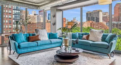 Stylish living room at AMLI 900 with teal sofas, round coffee table, exposed ductwork, and floor-to-ceiling windows framing sweeping Chicago skyline views