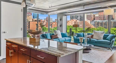 Open-concept kitchen and living room at AMLI 900 with granite island, pendant lighting, teal sofas, and walls of windows showcasing Chicago skyline views
