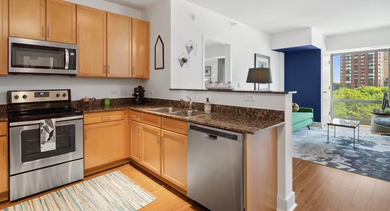 Modern kitchen at AMLI 900 featuring granite countertops, warm wood cabinetry, teal backsplash, and black appliances with a view toward the living area in Chicago