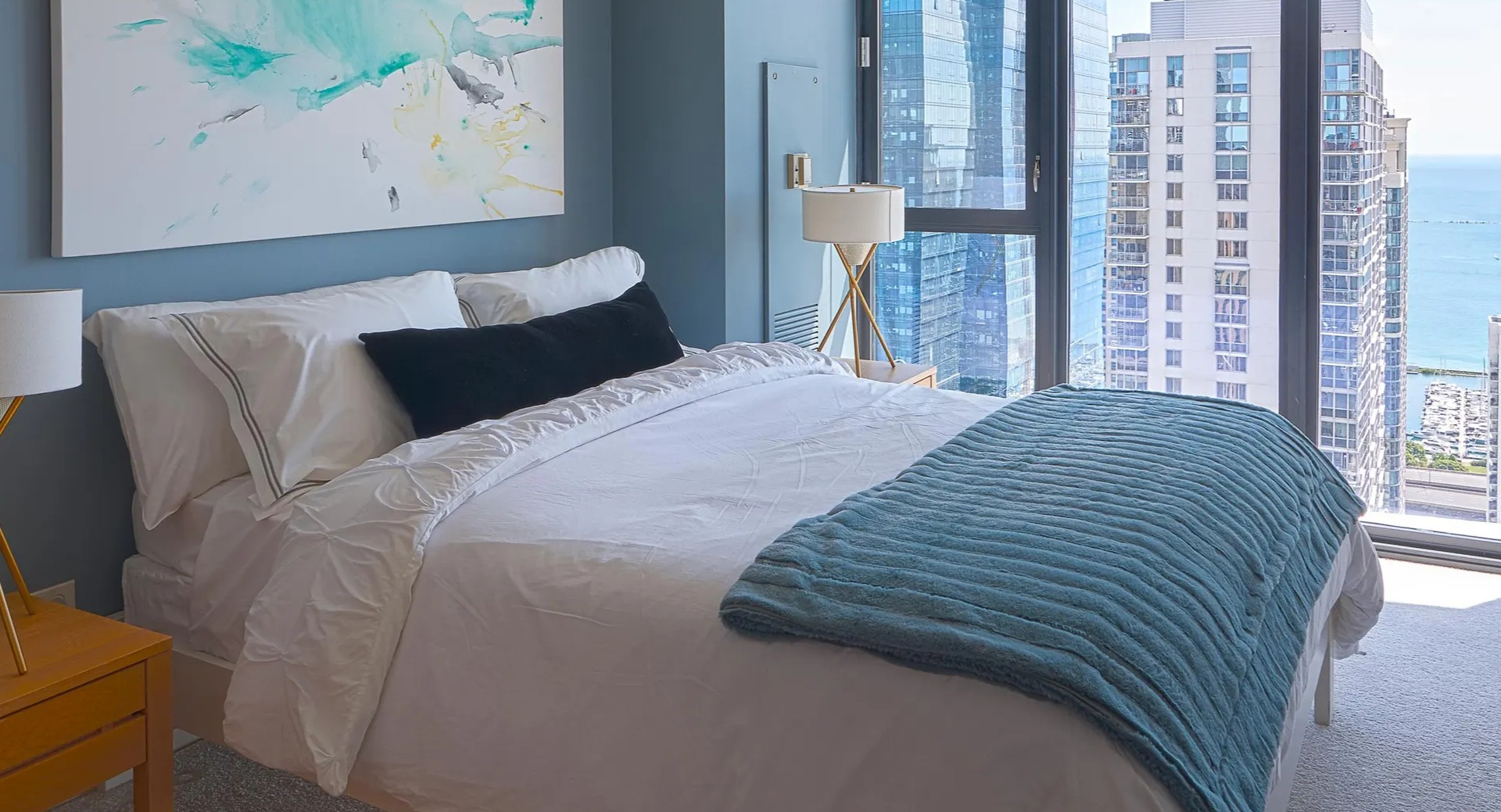 Bright primary bedroom with large windows, lake and skyline views, crisp white bedding, and a teal throw at Aqua at Lakeshore East Apartments in Chicago