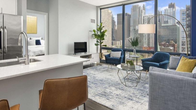 Spacious living room at Arkadia West Loop with large windows showcasing city views, leading to a cozy bedroom