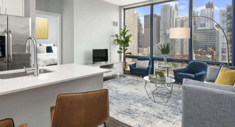Spacious living room at Arkadia West Loop with large windows showcasing city views, leading to a cozy bedroom