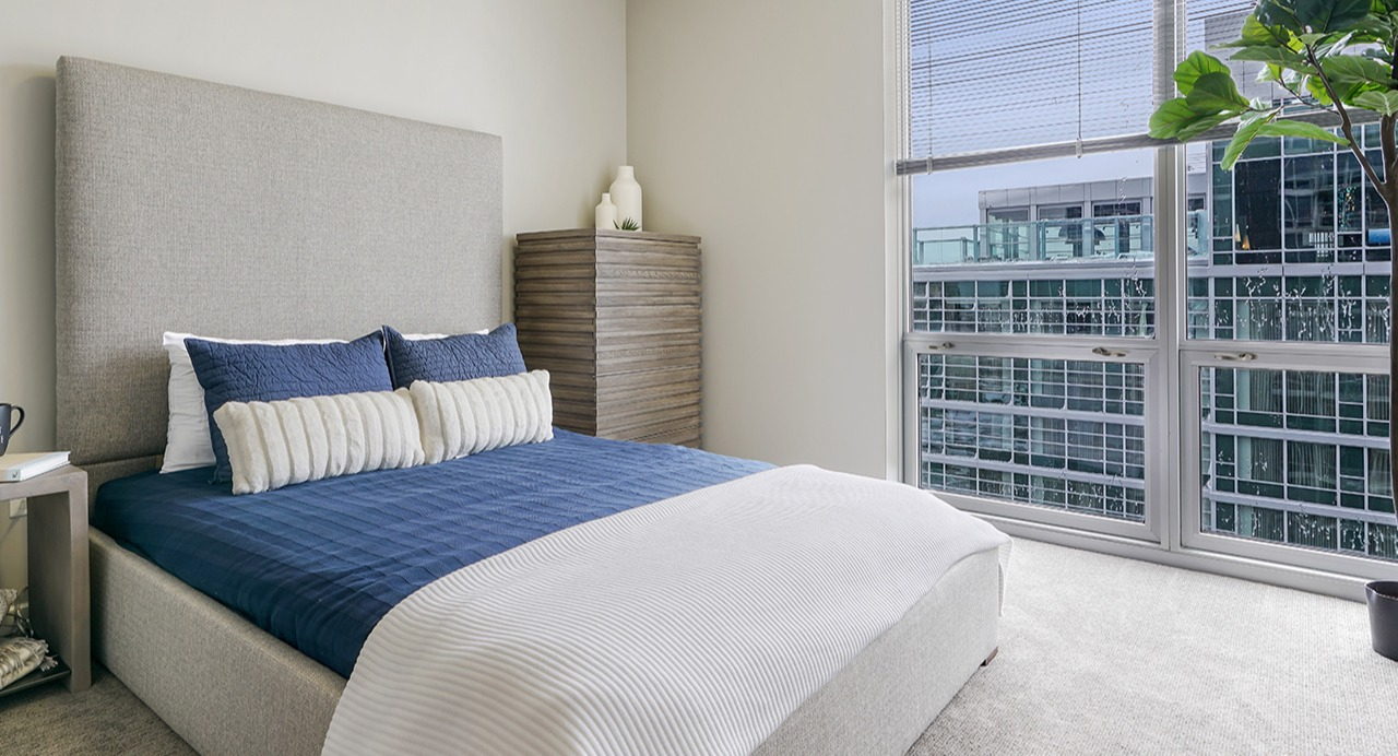 Cozy bedroom featuring a large window with city views, a comfortable bed with blue and white linens, and modern decor at Arrive LEX in Chicago
