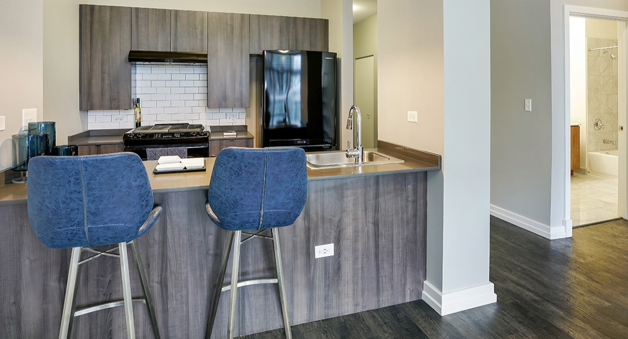 Modern kitchen with sleek appliances, wood cabinetry, and a breakfast bar, leading to a spacious hallway at Arrive LEX in Chicago