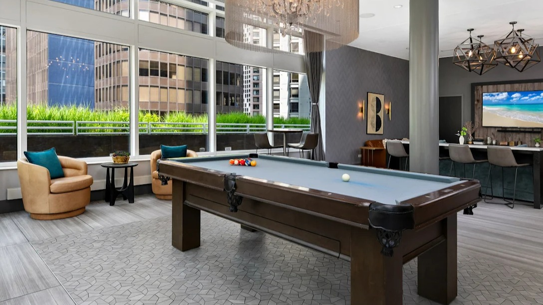 Stylish game room at Atwater Apartments in Chicago, featuring a billiards table, comfortable seating, and large windows with city views