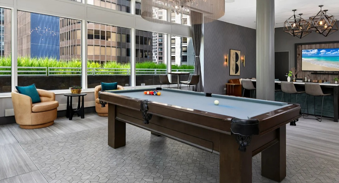 Stylish game room at Atwater Apartments in Chicago, featuring a billiards table, comfortable seating, and large windows with city views