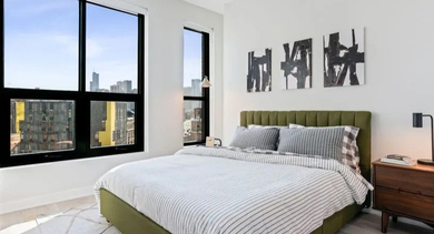 A cozy bedroom with a green upholstered bed, modern art, and large windows showcasing city views at Avenir apartments in Chicago