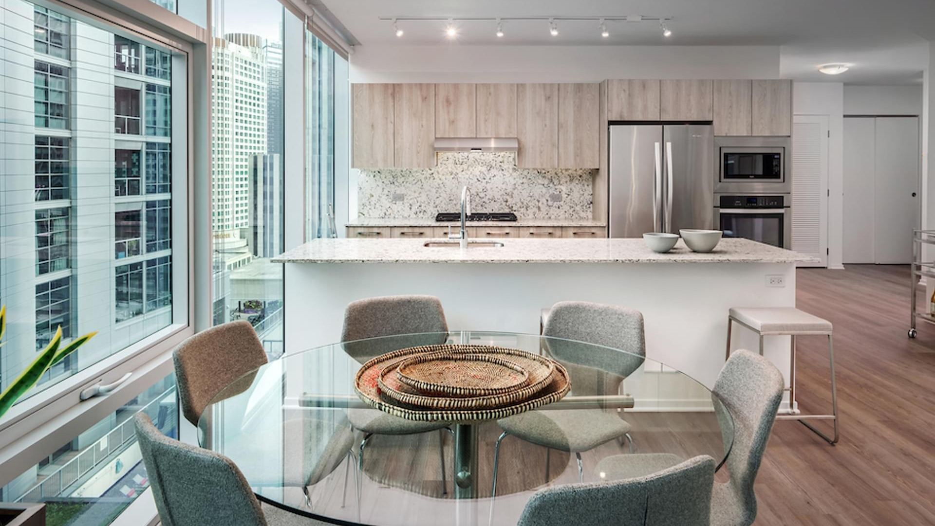 Bright dining area with round glass table, floor-to-ceiling windows, and a modern wood-grain kitchen at Optima Signature apartments in Chicago