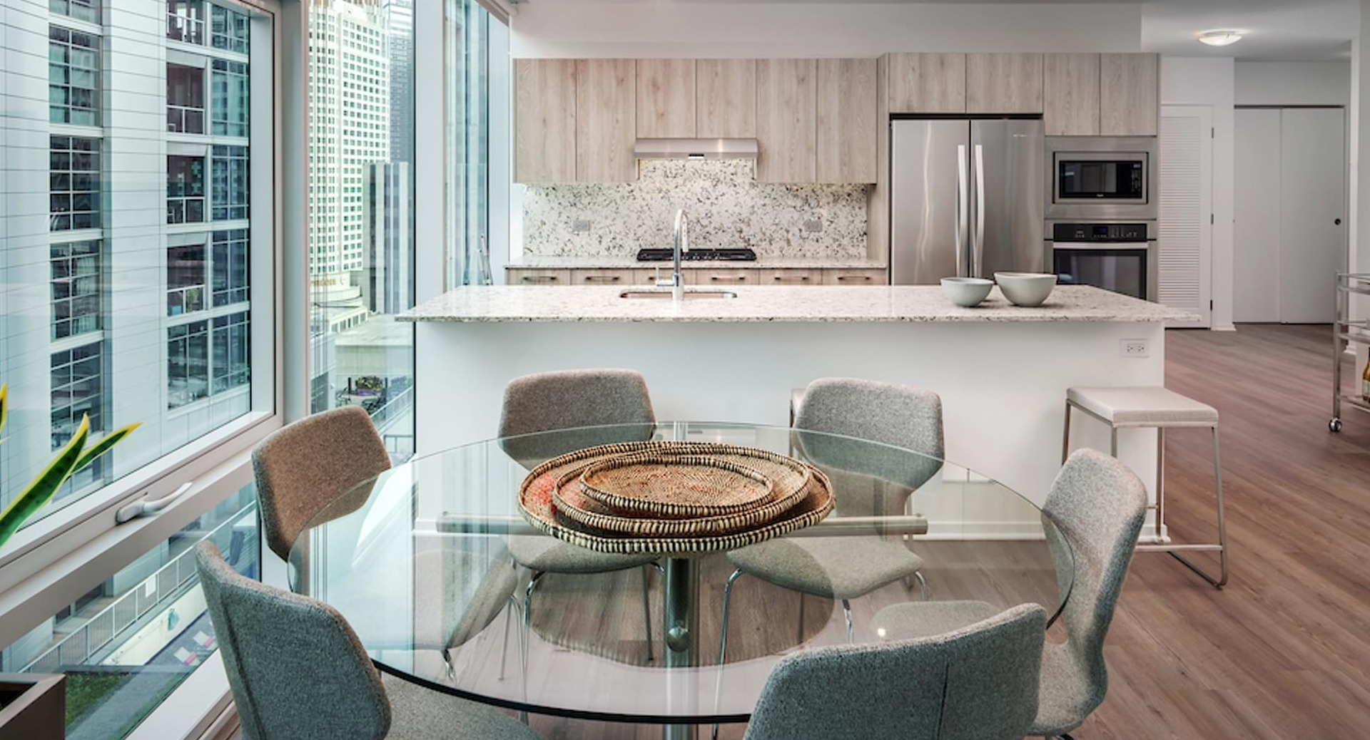Bright dining area with round glass table, floor-to-ceiling windows, and a modern wood-grain kitchen at Optima Signature apartments in Chicago