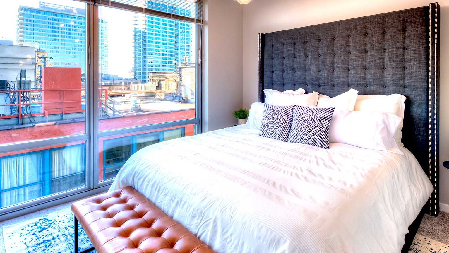 Cozy bedroom featuring a large bed with a tufted headboard, a stylish bench, and city views from expansive windows at Burnham Pointe in Chicago