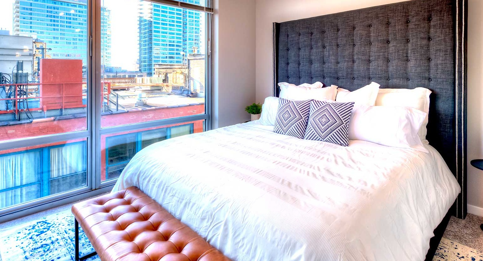 Cozy bedroom featuring a large bed with a tufted headboard, a stylish bench, and city views from expansive windows at Burnham Pointe in Chicago