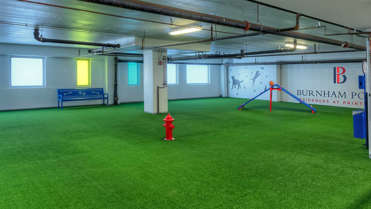 Spacious indoor dog park with artificial turf, a fire hydrant, and agility equipment, perfect for pets at Burnham Pointe in Chicago