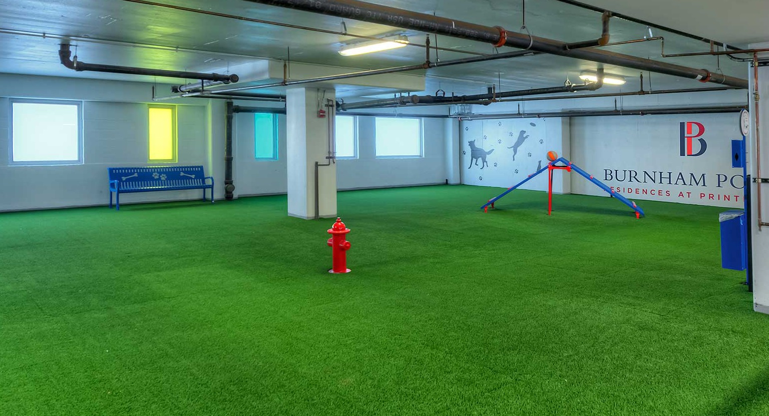Spacious indoor dog park with artificial turf, a fire hydrant, and agility equipment, perfect for pets at Burnham Pointe in Chicago