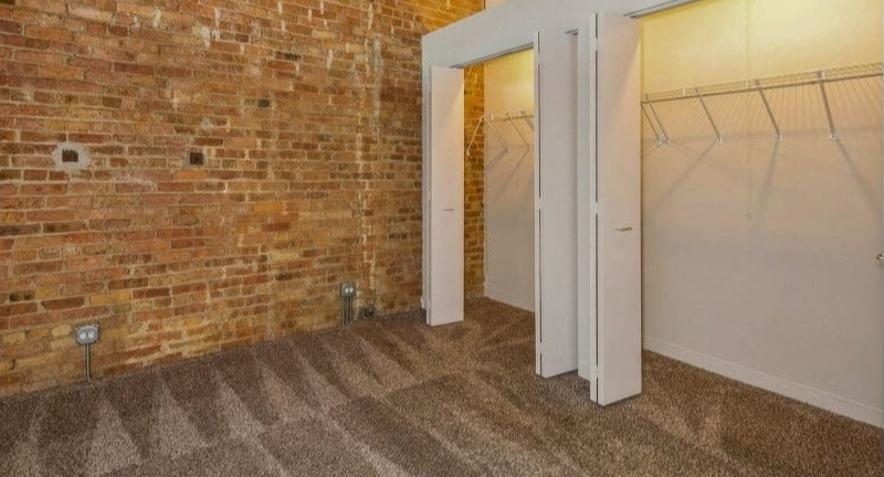 Bedroom space with an exposed brick wall and open walk-in closets, providing ample storage at Carriage House Lofts in Chicago