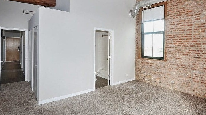 Cozy bedroom featuring soft carpet, exposed brick accent wall, and an ensuite bathroom entrance at Carriage House Lofts in Chicago