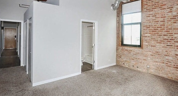 Cozy bedroom featuring soft carpet, exposed brick accent wall, and an ensuite bathroom entrance at Carriage House Lofts in Chicago