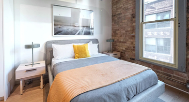 A cozy bedroom featuring exposed brick, a large window, and stylish modern furnishings at Cobbler Square Lofts in Chicago