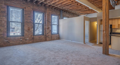 Cozy living room with exposed brick walls, large windows, and soft carpeting at Cobbler Square Lofts in Chicago