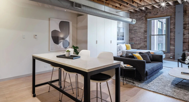 An open-concept studio apartment featuring a living area, dining table, and a bed with exposed brick at Cobbler Square Lofts in Chicago