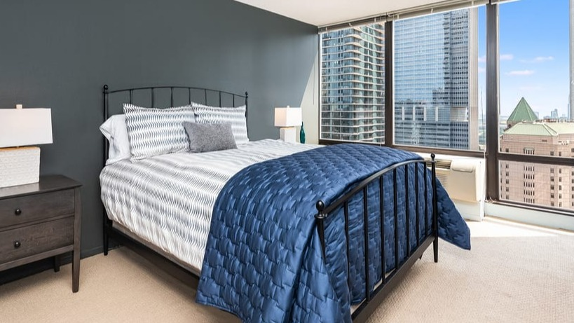 A comfortable bedroom with a dark bed frame, blue quilt, and large windows offering impressive city views
