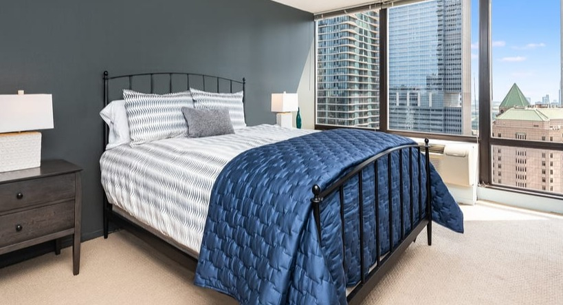 A comfortable bedroom with a dark bed frame, blue quilt, and large windows offering impressive city views