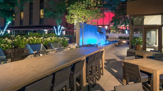 A beautifully lit outdoor patio at dusk, featuring dining tables, grills, and a glowing water feature