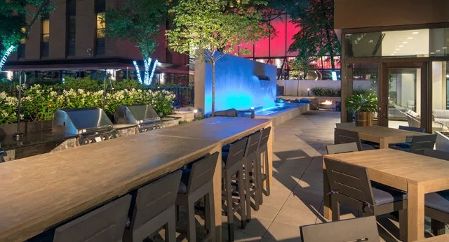  A beautifully lit outdoor patio at dusk, featuring dining tables, grills, and a glowing water feature