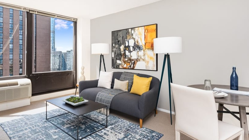 A contemporary living room with a grey sofa, colorful accents, dining area, and large windows with city views