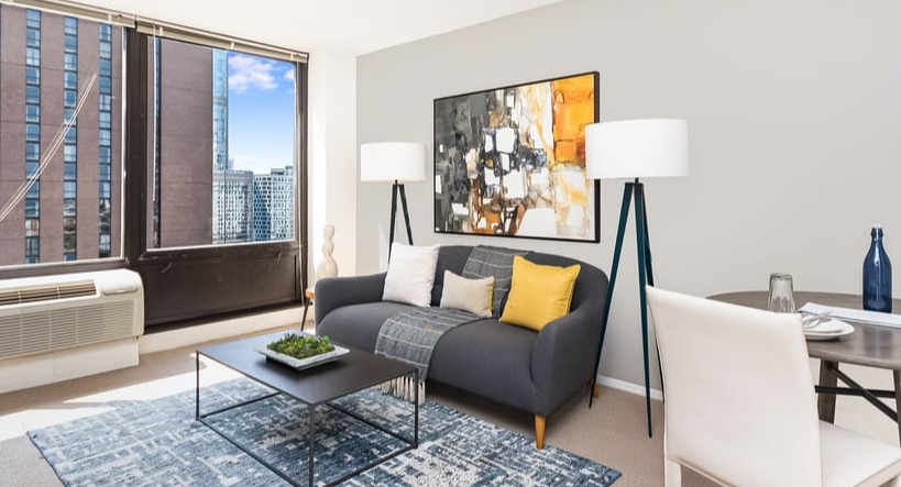 A contemporary living room with a grey sofa, colorful accents, dining area, and large windows with city views