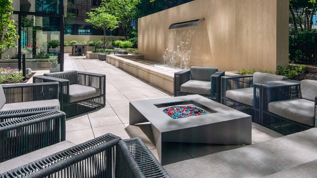 A modern outdoor lounge area with a fire pit, comfortable seating, and a water feature in a lush setting