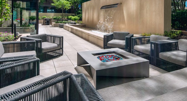 A modern outdoor lounge area with a fire pit, comfortable seating, and a water feature in a lush setting