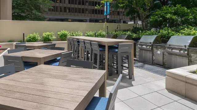 A spacious outdoor area with dining tables and multiple gas grills, surrounded by greenery and city buildings