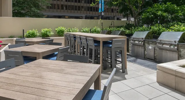 A spacious outdoor area with dining tables and multiple gas grills, surrounded by greenery and city buildings