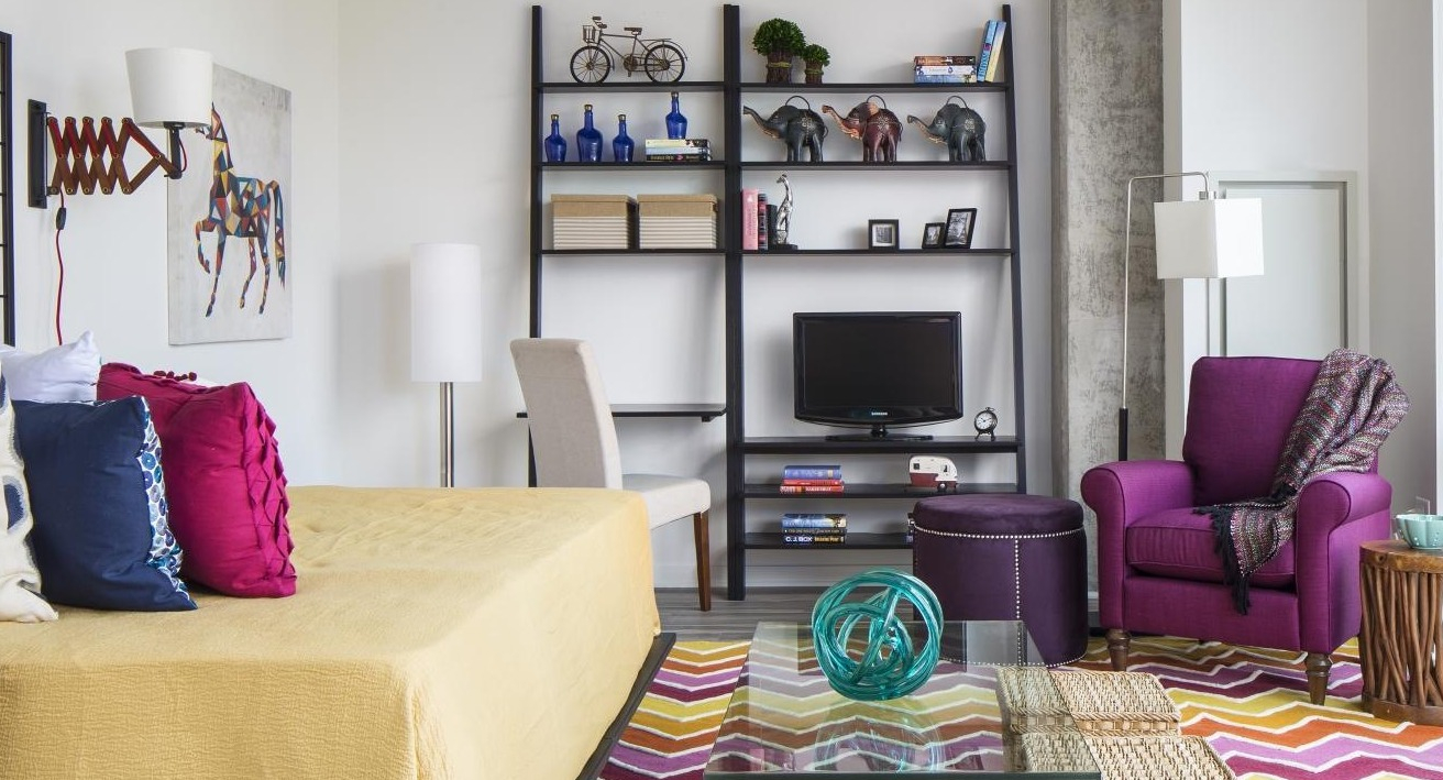 Vibrant studio apartment bedroom with a yellow bed, purple armchair, and colorful rug at Eight O Five Apartments in Chicago