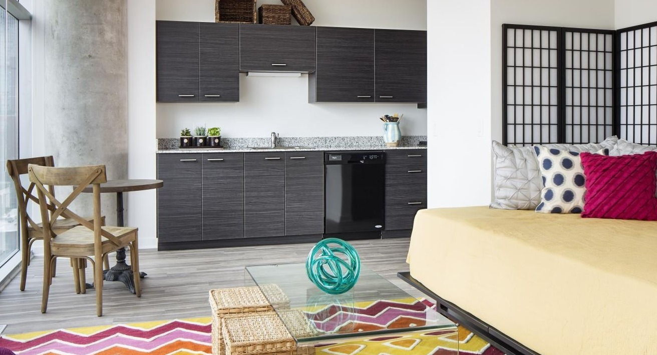 Compact studio apartment with a dark modern kitchenette, vibrant rug, and bed with colorful pillows at Eight O Five Apartments in Chicago