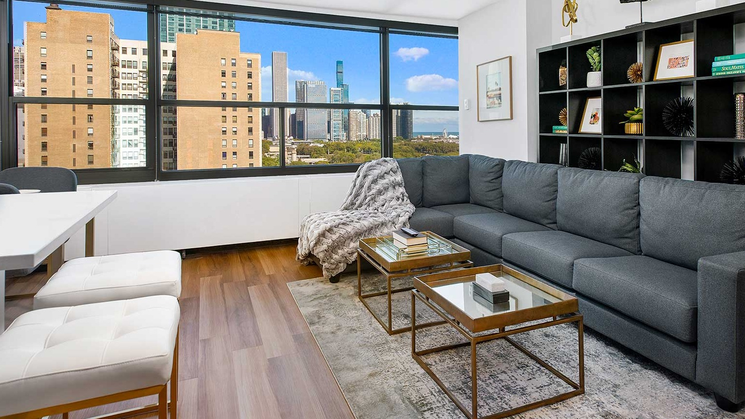 Elegant living room featuring a large sectional sofa, shelving, and expansive windows offering panoramic city views at Eleven Thirty in Chicago