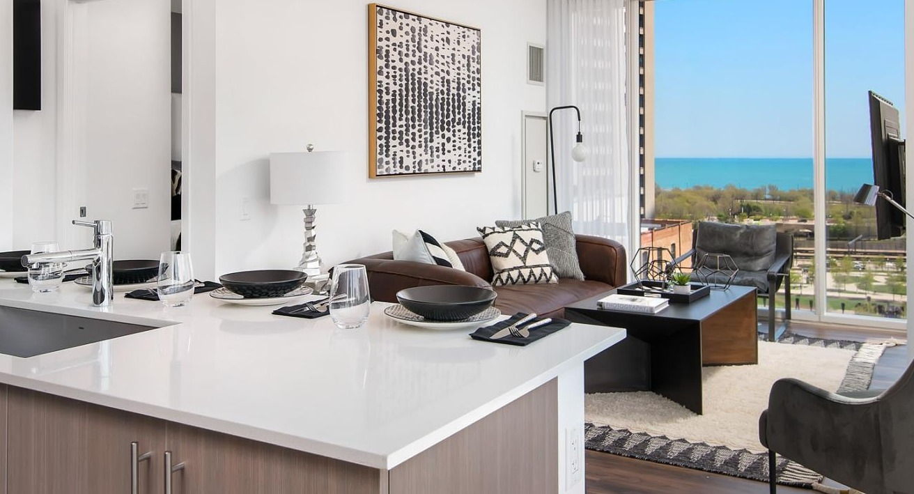 Modern apartment kitchen with a dining island, flowing into a living room with expansive windows showcasing lake views at Eleven40 in Chicago
