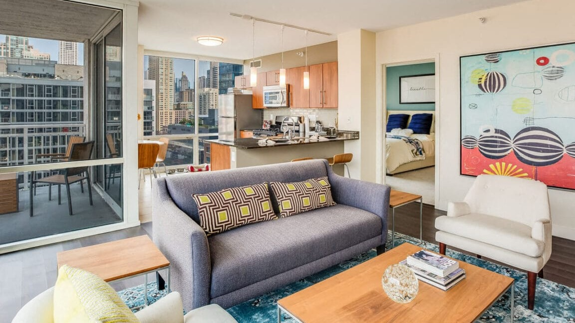 Open-concept apartment living room at Flair Tower Apartments in Chicago, showcasing the adjacent kitchen, balcony, and a glimpse into the bedroom
