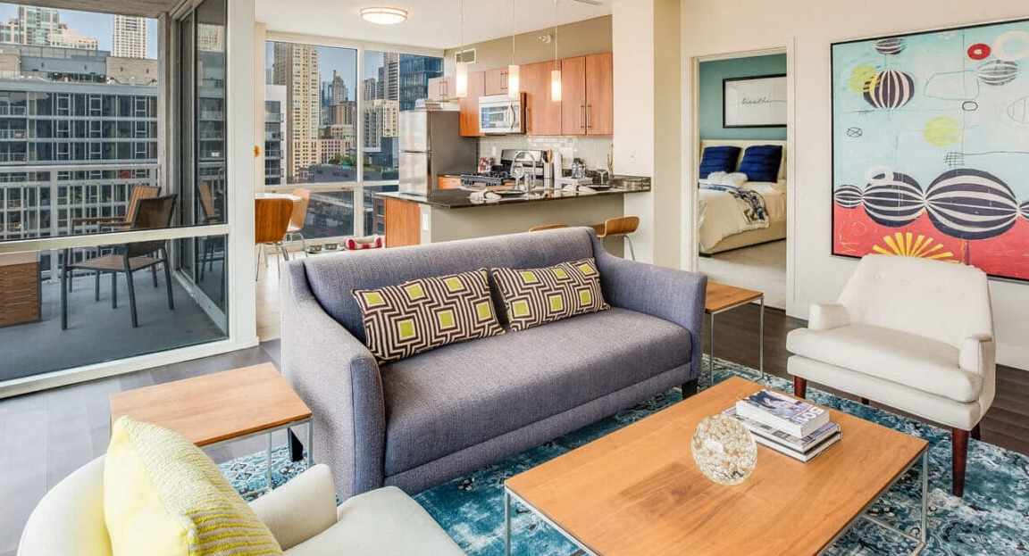 Open-concept apartment living room at Flair Tower Apartments in Chicago, showcasing the adjacent kitchen, balcony, and a glimpse into the bedroom