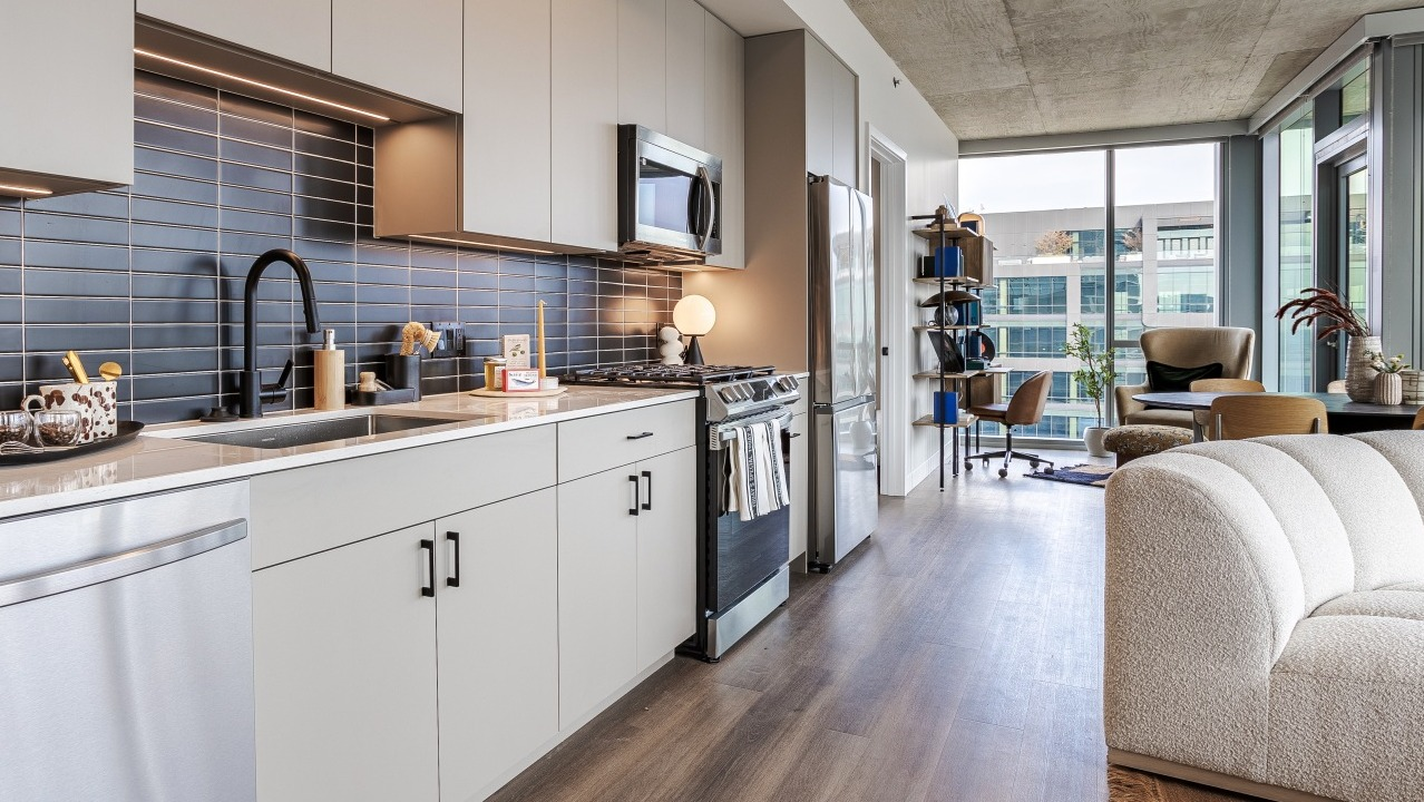Bright apartment kitchen at Flora in Chicago, featuring light cabinetry, a dark subway tile backsplash, and an open view into the living space