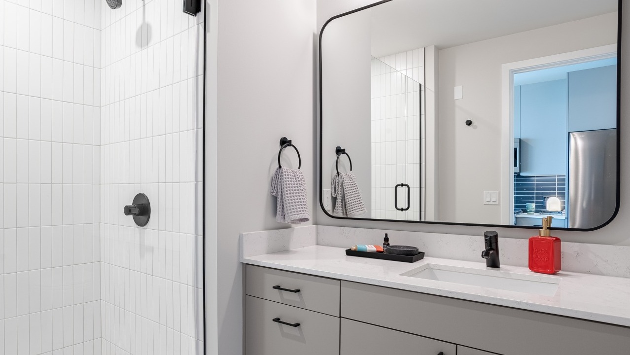 Sleek, modern apartment bathroom at Flora in Chicago, featuring a grey vanity, stylish mirror, and a spacious walk-in shower with subway tiles