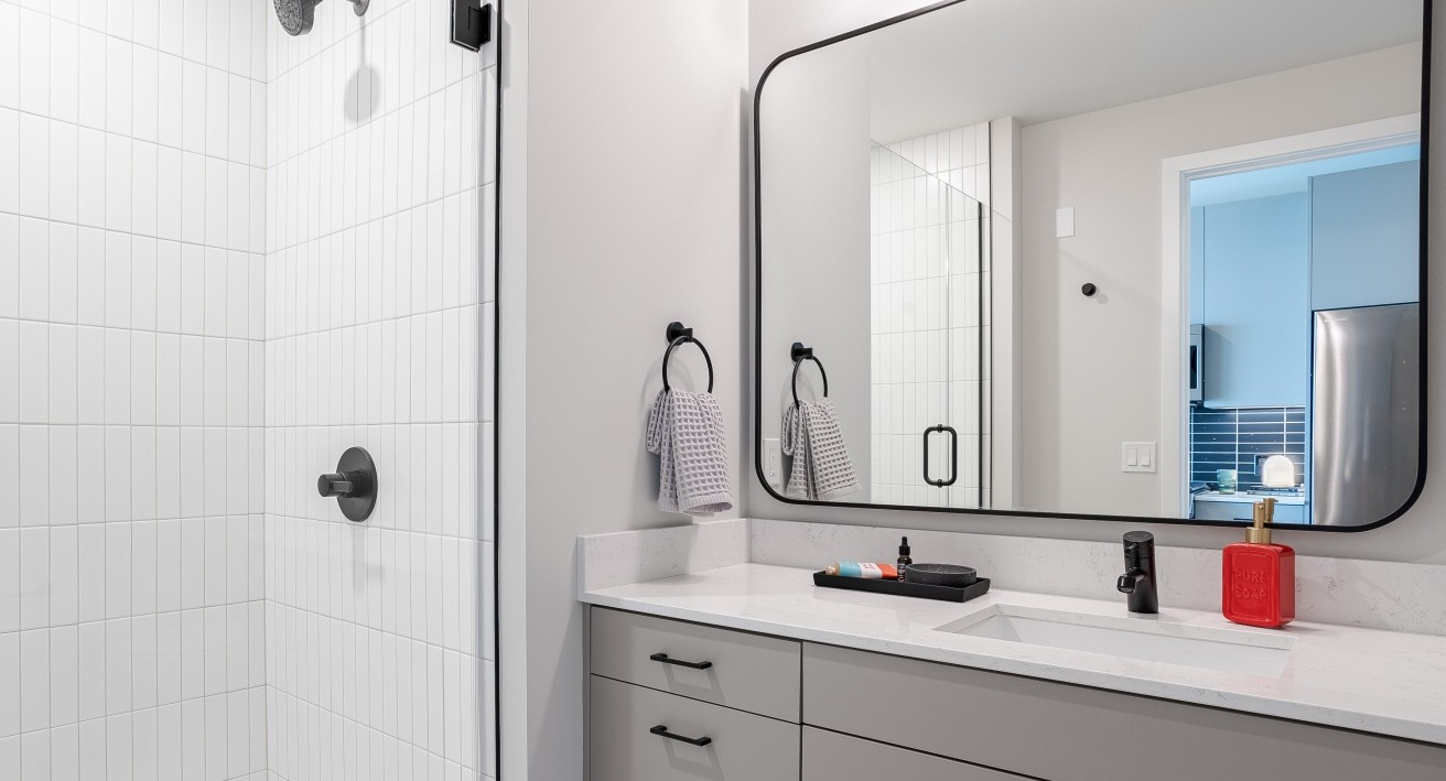 Sleek, modern apartment bathroom at Flora in Chicago, featuring a grey vanity, stylish mirror, and a spacious walk-in shower with subway tiles