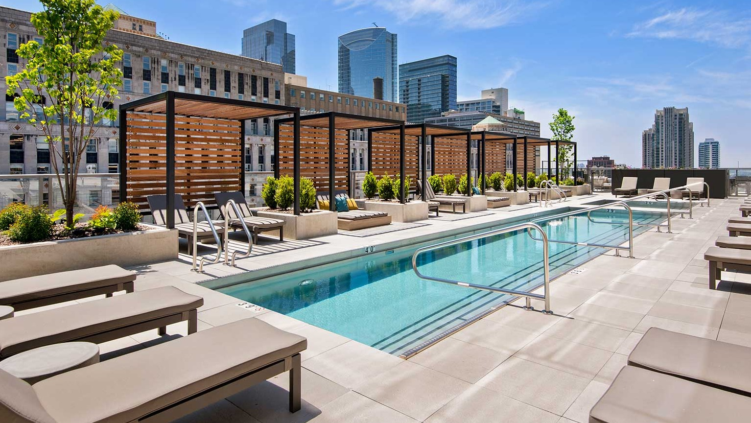 A serene outdoor pool area at Hubbard 221 with numerous lounge chairs, pergolas, and city buildings in the background
