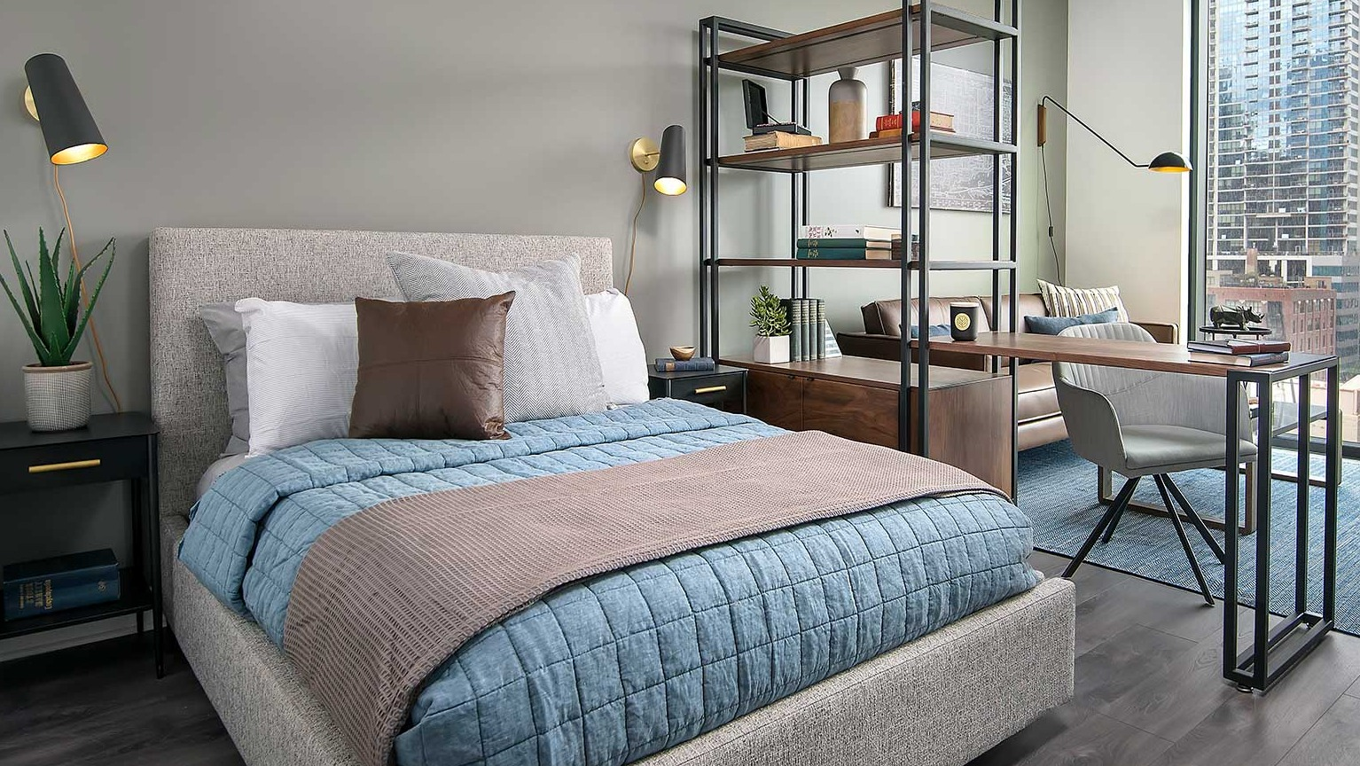 A well-designed studio apartment at Hubbard 221, showcasing a bed, work area, and city views from large windows