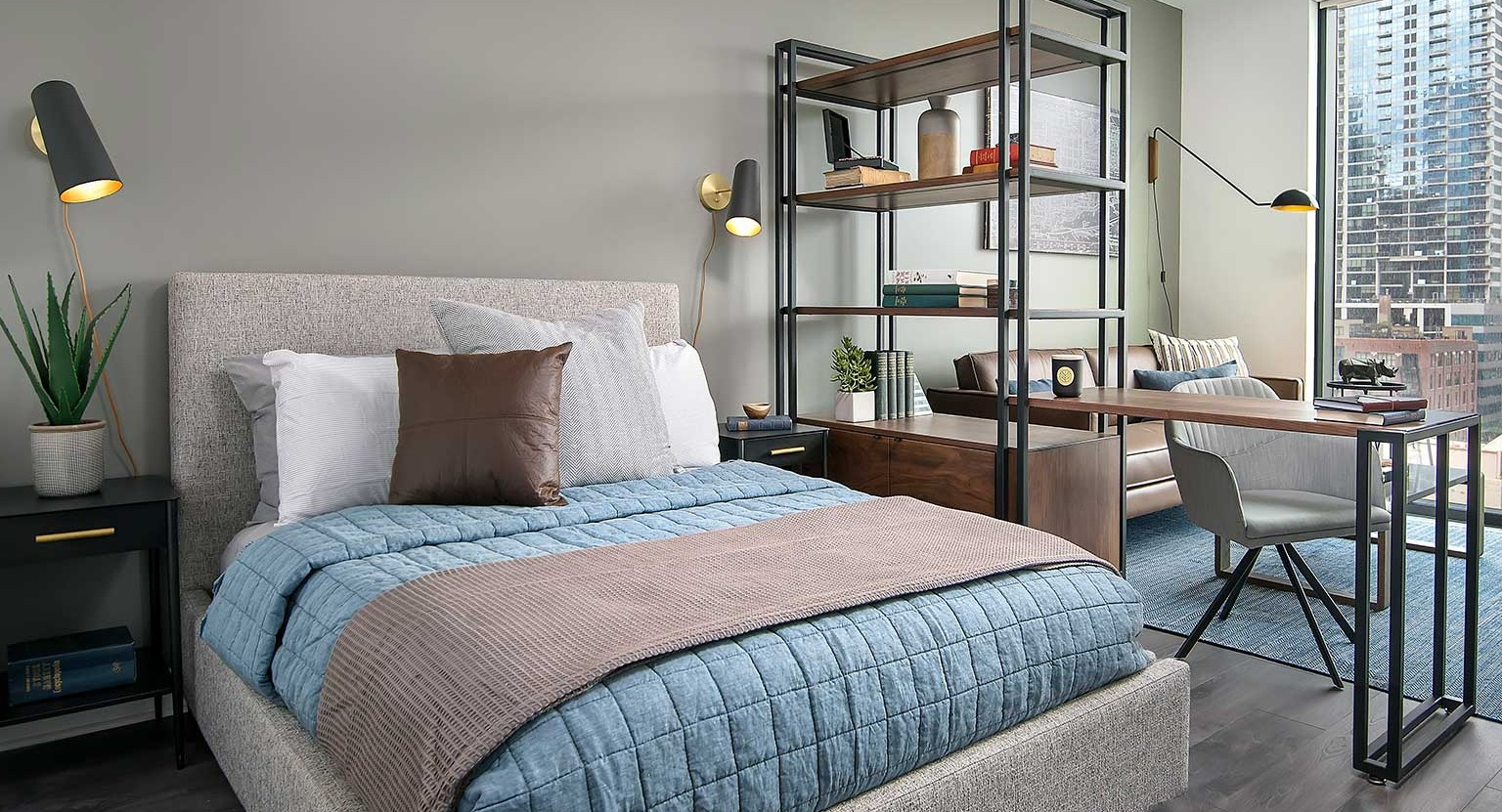 A well-designed studio apartment at Hubbard 221, showcasing a bed, work area, and city views from large windows