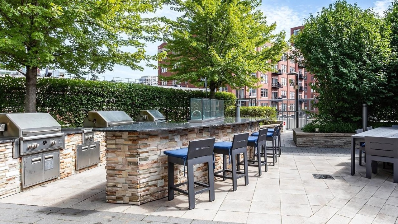 Outdoor grilling station with multiple barbecues and an extended bar seating area, surrounded by greenery at Hubbard Place in Chicago