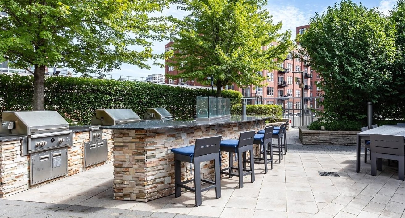 Outdoor grilling station with multiple barbecues and an extended bar seating area, surrounded by greenery at Hubbard Place in Chicago